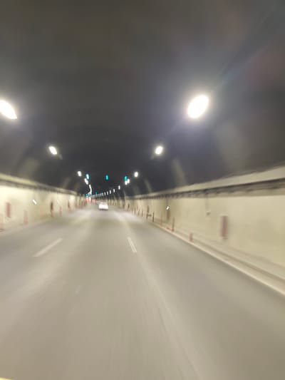 This is a blurry, motion-captured image of a vehicle's perspective traveling through a tunnel. The tunnel has a dark, arched ceiling with overhead lights that are visible as bright, elongated streaks due to the motion. The tunnel walls are a light color, possibly concrete, and are lined with what appear to be safety features like red-topped bollards and red rectangular emergency boxes. There are traffic lane markings visible on the asphalt road. In the distance, a white car is visible, also blurred by motion. Green traffic lights are also visible overhead. The image appears to have been taken during daylight hours as indicated by the brightness of the lights in the tunnel. The scene is devoid of any discernible people. The provided location of Gorno Kamartsi, Bulgaria, cannot be verified or denied based solely on the visual information in the image, as there are no identifying landmarks or textual elements present.