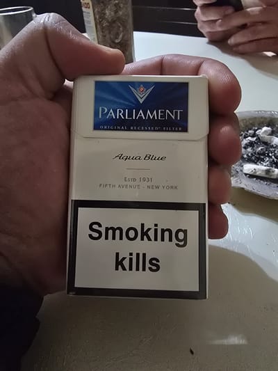 A hand is holding a pack of Parliament Aqua Blue cigarettes. The pack has a blue and white design with the Parliament logo and "ORIGINAL RECESSED FILTER" printed on the top portion. Below that, it says "Aqua Blue" in cursive script. Further down, "ESTD 1931" and "FIFTH AVENUE - NEW YORK" are printed. The bottom of the pack features a black and white graphic with the text "Smoking kills" in large, bold letters. In the background, there appears to be a blurred person's hands and a jar of what looks like loose tobacco. To the right of the cigarette pack, a small ashtray with cigarette butts is visible. The scene is lit by overhead lighting, casting shadows on the table surface. The location in the city of Khasavyurt, Russia is not discernible from the image.