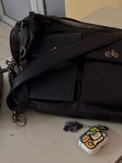 This image depicts an indoor scene in the city of Brčko, Bosnia and Herzegovina. The primary subject is a black, textured synthetic leather shoulder bag, positioned on a light-colored, flat surface. The main compartment of the bag is partially open, revealing a metallic zipper track. A wide, black shoulder strap diagonally crosses the front of the bag. Two front flap pockets are visible, each secured by a silver-colored metallic twist-lock clasp; one clasp is more distinctly visible. On the left side of the bag, a silver-colored chain is attached to a ring, from which a small, silver-colored, heart-shaped padlock hangs.
On the surface immediately in front of the bag, two keychains/charms are present. One is a white wireless earbud charging case, adorned with colorful cartoon illustrations of multiple cats. A black carabiner-style clip is attached to this case. Adjacent to it, and possibly connected, is a smaller, transparent rectangular charm featuring a cartoon depiction of a grey dog's head.
The light-colored surface on which these items rest shows minor scuffs or dust. The background is out of focus, revealing a muted yellowish-beige wall and a segment of a light grey or metallic doorframe or window frame in the upper left. No individuals are present in the image, and no specific actions or interactions are depicted.