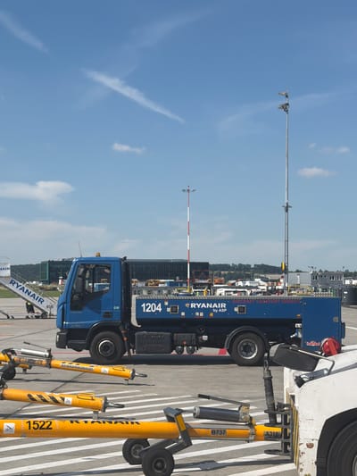 The image shows an airport tarmac at John Paul II International Airport Kraków–Balice in Balice, Poland. In the foreground, there are several yellow tugger bars, one with the number "1522" and "RYANAIR" printed on it. Behind these, a blue truck with "RYANAIR" and "AIRCRAFT LAVATORY SERVICE" written on its side is parked. The truck has the number "1204" visible on its tank. To the left of the truck, a mobile stairway with "RYANAIR" branding is positioned near an unseen aircraft. In the background, a modern terminal building with a dark facade and a row of trees can be seen under a clear blue sky with some wispy clouds. A tall pole with lights is also visible.