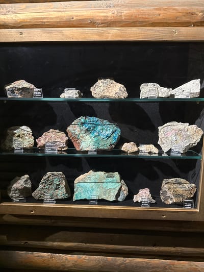 The image shows a display of various mineral specimens in a wooden cabinet with glass shelves. The cabinet is positioned in what appears to be a museum or collection space, with a dark background behind the shelves. The mineral specimens vary in size, color, and texture. Some are rough and earthy, while others exhibit vibrant blue and green hues, suggesting copper-based minerals. Each specimen has a small, clear label in front of it, indicating its name and origin. One of the labels reads "Grube Anton im Heubach b. Schil-t ach," which, when combined with the context of minerals, strongly suggests the display is located in or related to the area of Schiltach, a town near Schonach im Schwarzwald, Germany, given the geological context implied by the labels. There are no people visible in the image. The lighting appears to be artificial, highlighting the specimens on display.