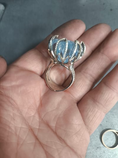 A close-up, top-down, angled shot shows a person's open palm, with a large, ornate ring resting on it. The ring features a prominent, faceted, light blue gemstone, possibly aquamarine, that appears to be carved in a floral or crown-like design. The setting of the ring is silver or white gold, encrusted with small, clear stones, likely diamonds, along the band and the prongs holding the main gemstone. The hand shows visible lines and texture, with a slight dusting of what appears to be fine particulate matter on the skin and the ring setting. In the lower right corner of the frame, another plain gold ring is visible on a grey, textured surface. The background is a muted, greyish surface. There is no indication of the city of Sredneuralsk, Russia, or any other specific geographical location.