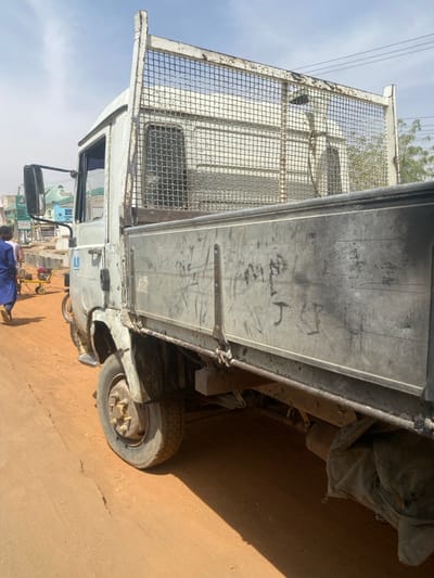 The image displays the driver's side of a weathered, light grey flatbed truck parked on an unpaved, reddish-brown dirt road in Bachirawa, Nigeria. The truck exhibits extensive dirt, rust, and faded paint. Its metal flatbed features tall, dirty side panels with multiple scratches and black, indistinct markings. A rusted metal mesh guard extends upwards from the front of the bed, behind the cab. The visible front wheel shows a worn tire and rusted hub. Beneath the truck bed, undercarriage components and a dark, possibly tarp-covered object are visible.
To the left of the truck, a person wearing a blue garment and light-colored shoes is seen walking away from the viewer. Near this person, a small yellow wheeled cart or piece of equipment is situated on the dirt road. In the background, several buildings, including one with a green roof and a sign partially reading "MAMA," line the roadside. Sparse green vegetation and overhead utility lines are visible in the distance against a clear, light blue sky with minimal clouds. The scene is illuminated by bright daylight, casting shadows beneath the truck and other objects.