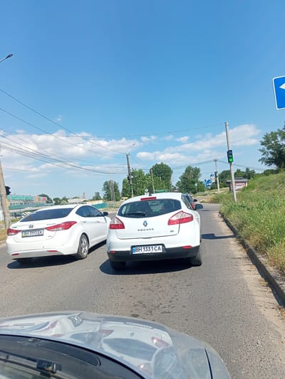 This media file depicts a view from inside a vehicle traveling on a paved road under a bright blue sky with scattered clouds. The scene is set during the daytime, likely late morning or early afternoon, given the strong sunlight casting shadows.

The primary subjects are two white cars: a Hyundai Elantra in the left lane and a Renault Megane in the right lane, ahead of the camera's vehicle. Both cars are facing forward, suggesting they are in traffic. The Hyundai Elantra has a license plate visible that reads "BH 3993 EK" and "ODESSA". The Renault Megane has a license plate that reads "BH 1331 CA".

Other objects in the scene include a traffic light displaying a green signal, power lines crisscrossing the sky, and streetlights. In the background, there are trees and buildings, including what appears to be a church with blue domes. The right side of the road is bordered by a grassy embankment with weeds and some dry vegetation.

A blue traffic sign with a white arrow pointing left is visible in the upper right portion of the frame. The overall impression is a typical day on a suburban or semi-rural road, possibly in Bilhorod-Dnistrovskyi, Ukraine, as indicated by the location context. There are no people visible in the frame, and the mood is neutral, simply documenting a moment of travel.