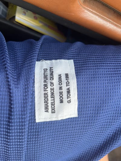 The image shows a close-up of a blue, textured fabric with a white label attached. The label has black text printed on it. The text reads: "AWARDER FOR PURITYG EXCELLENCE OF QUAUTY MOOE IN CGINA G.TOMA TO-1998". The fabric appears to be a garment, possibly a shirt or jacket. The background is blurred, but suggests an indoor setting with some objects in the distance, possibly a brown leather surface and a yellow item with some writing. There is no visible indication of the city of Aqköl, Kazakhstan, or any specific location within it.