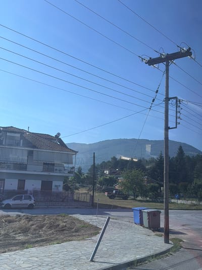 The image depicts an outdoor scene, likely in Kato Nevrokopi, Greece. The primary subjects are a utility pole with numerous wires stretching across the frame against a clear blue sky. In the middle ground, a multi-story building with a tiled roof and balconies is visible. A white car is parked in front of the building. The background is dominated by a hazy, tree-covered mountain.
To the right of the building, a paved area with a stone pattern is visible, bordering a patch of dry grass or weeds. A large blue dumpster with green and white stripes is also present in this area, next to a utility box. A metal pole, possibly a street lamp or signpost, stands in the foreground on the right.
The time of day appears to be daytime, with bright sunlight casting shadows. The weather seems clear and sunny. No people are clearly visible in the image. The overall scene suggests a residential or semi-rural setting with infrastructure for electricity and waste management. There are no discernible text elements in the image.