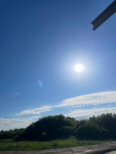The image shows a bright, sunny day with a clear blue sky. The sun is high in the sky, casting a bright glare. A dark, angular object is visible in the upper right corner of the frame, possibly a piece of construction material or a piece of equipment. Below the sky is a dense line of green trees, forming a natural horizon. In the foreground, there is a patch of dry, dusty ground, likely a road or construction site, with scattered small rocks and debris. The overall scene suggests an outdoor, possibly rural or semi-constructed environment, consistent with the outskirts of a settlement like Сокуры, Russia, on a clear day.