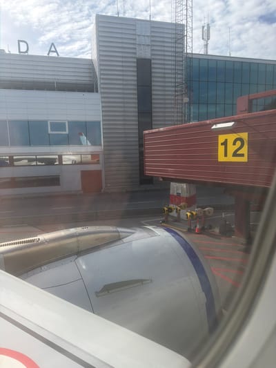 This is an exterior, eye-level, medium shot of a passenger jet engine and wing seen through an airplane window. The view is from inside the aircraft looking out at an airport terminal building with a jet bridge attached. The terminal building has large glass windows and a metallic facade. The jet bridge is red with the number "12" prominently displayed in yellow on its side. Various airport equipment, including yellow and red chocks and cones, are visible on the tarmac below the jet bridge. The sky is partly cloudy. The image appears to be taken at Stockholm-Arlanda Airport (ARN) in Stockholm, Sweden, based on the presence of the "DA" signage on the terminal building which is consistent with Arlanda's airport code.