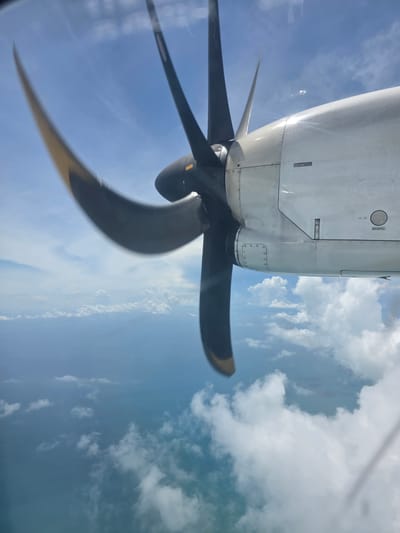 The image captures a close-up view of a propeller and engine nacelle of an aircraft, seen from inside the cabin through a window. The propeller blades, painted dark with yellow tips, are blurred, indicating motion, and extend across the frame against a backdrop of a bright blue sky dotted with white, fluffy clouds. The engine nacelle is a clean, white structure with visible rivets and panel lines, suggesting it is part of a turboprop aircraft. The perspective is from an elevated viewpoint, looking downwards and outwards.
The lighting suggests it is daytime, likely mid-morning or afternoon, given the brightness of the sky and the sunlight reflecting off the aircraft's surface. The weather appears clear with scattered clouds, typical of a tropical climate. While the exact location is not explicitly visible, the context provided points to Ban Tai, Thailand. There are no people or specific emotional cues evident in the image. The scene depicts an airplane in flight. A small, rectangular label with indistinct text is visible on the engine nacelle, but the text is not legible.