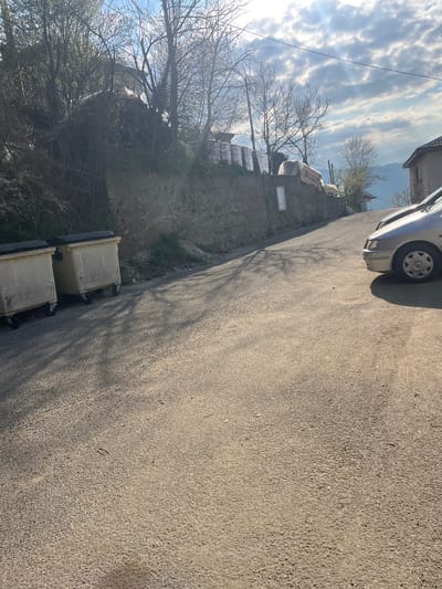 The image depicts a street scene in Churka, Bulgaria. The foreground is dominated by an asphalt road. A portion of a silver car is visible in the top right corner. Along the left side of the road is a low stone wall. Behind the wall is overgrown vegetation and bare trees. Two yellow and black trash bins are located on the left side of the road near the wall. In the background, glimpses of buildings and a cloudy sky can be seen. The overall lighting is bright with shadows cast by the trees.