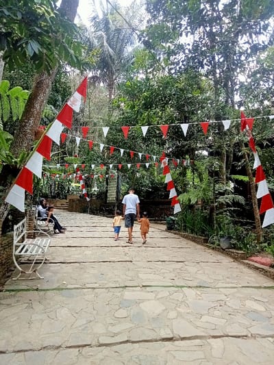 The image depicts an outdoor recreational area in Kajar, Indonesia, characterized by a wide, ascending stone-slab path. Red and white Indonesian flags and triangular pennants are extensively strung across and along the path, indicating a festive or patriotic display.

On the right side of the path, a male adult, viewed from behind, wearing a white t-shirt and dark shorts, walks uphill with two small children. One child, to his left, wears a yellow top and blue shorts; the other, to his right, wears an orange or brown top and shorts. All three are moving away from the viewer.

On the left, a white ornate metal bench is positioned beside the path. A woman, wearing a dark head covering and a light-colored long-sleeved garment, is seated on this bench, accompanied by another individual, possibly a child, partially obscured.

The environment is lush and verdant, with dense green foliage, tall trees, and ferns lining both sides of the path. Palm trees are visible in the background. The scene is illuminated by natural daylight, suggesting an outdoor setting.