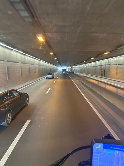 The image depicts a view from inside a vehicle traveling through a tunnel. The tunnel's ceiling is made of concrete with visible expansion joints and rows of lights. The walls of the tunnel are lined with horizontal panels. On the road, there are several vehicles. In the foreground, a dark-colored car is visible on the left lane. Further ahead, a silver car is in the right lane, and a white truck is in the left lane. The road has clear white lane markings. In the lower right corner, part of a GPS device is visible. The environment suggests an urban setting, possibly a major thoroughfare within a city. However, no specific landmarks are identifiable that would definitively place the location in Augsburg, Germany.