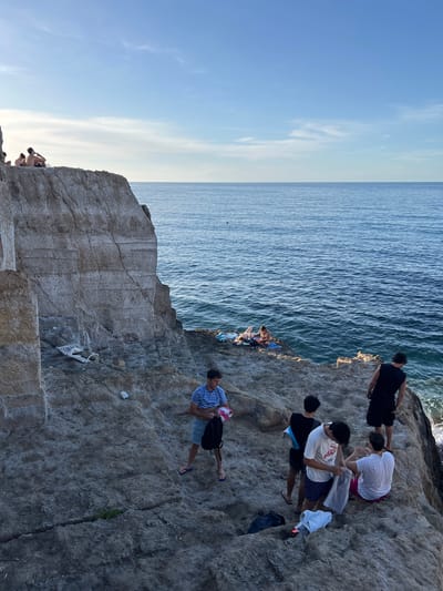 This image depicts a coastal scene in Sperone, Italy, under clear blue skies, likely in the late afternoon. The foreground features a large, light-colored stratified rock cliff on the left, with rough, uneven rocky ground extending towards the right and the sea.
On the upper left edge of the main cliff, two individuals are seated, observing the distant horizon. Further down the cliff face, resting on a small ledge, is an overturned and broken white plastic garden chair, accompanied by scattered small pieces of white litter.
The deep blue sea occupies the right side of the frame, showing visible ripples on its surface. On a lower rocky outcrop by the water, two individuals are relaxing on blue and white towels. One person is reclining, partially obscured by a towel, while another sits upright.
In the lower right section of the image, a group of approximately seven young men is gathered on the rocky terrain. One man in a blue and white striped t-shirt and patterned shorts, wearing blue flip-flops, stands holding a black bag and a pink item. Closer to the water's edge, a man in a black tank top and shorts stands looking out. Another man in a black tank top stands facing inwards. A third man in a white t-shirt with a graphic and patterned shorts is seen holding a grey garment from a clear plastic bag. Seated nearby is a man in a white t-shirt and pink/red shorts, looking towards the individual with the bag. Scattered on the rocks around this group are a black bag, a white towel, and other personal items in plastic bags. A small patch of green vegetation grows in a crack in the rock at the bottom center.