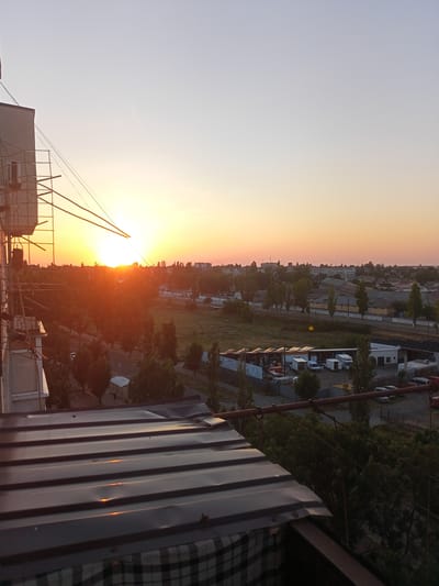 This media file captures a sunset over the city of Bilhorod-Dnistrovskyi, Ukraine. The scene is viewed from an elevated position, likely a balcony or rooftop, as indicated by the corrugated metal roof in the foreground and the side of a building with scaffolding.

The sun, a bright, intense orb, is low on the horizon, casting a warm, orange-yellow glow across the sky and the landscape. The light flares dramatically, creating lens flare effects and silhouetting the trees and buildings in the mid-ground. The sky transitions from a pale yellow near the sun to a soft, clear blue at the top of the frame, suggesting fair weather.

Below the sunset, the cityscape unfolds. Rows of trees line streets and form green swathes across the land. Various buildings, including what appear to be industrial or commercial structures with long, low roofs, and residential buildings, are visible. Some white vans and other vehicles are parked in an open area with solar panels, possibly indicating a business or industrial park. The overall impression is one of a typical urban and semi-industrial environment bathed in the serene light of sunset.

The time of day is clearly sunset, characterized by the low sun angle and the warm, vibrant colors in the sky and on the landscape. The location context of Bilhorod-Dnistrovskyi, Ukraine, is provided externally. There is no visible text within the image. The scene evokes a sense of calm and the peaceful end of a day.