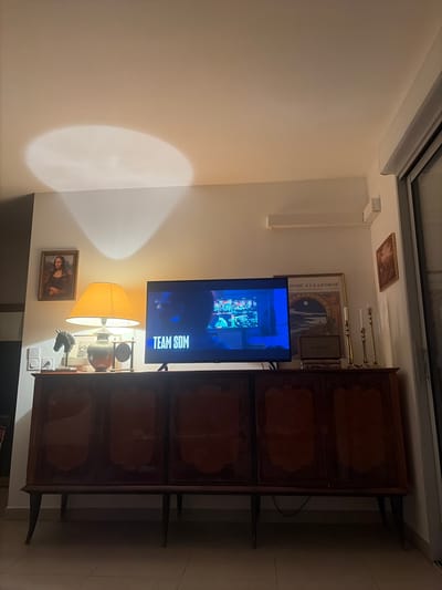 A wall displays a framed Mona Lisa print, a table lamp, a television, a framed art print, and a wall-mounted light fixture. Above the television, there is a framed picture and candelabras. To the right of the television is a dark-wood cabinet with decorative panels. A bright light shines on the adjacent wall. The image appears to be rotated slightly, presenting a side view of the wall's features.
