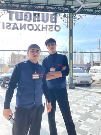 Two men wearing blue vests, black turtlenecks, and matching beanies stand side-by-side, each with a name tag that reads "BURCUT OSHXONA." The sign for "BURCUT OSHXONA" is visible in the background. One man holds a black object in his hand, possibly a remote or a phone. The photo is taken at an angle, with a tiled floor pattern visible. Cars can be seen parked in the background, including a white van and a gray SUV.