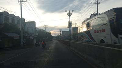 The image shows a street scene in Geulangganggampong, Indonesia, during what appears to be the late afternoon or early morning given the lighting. A white bus with "Kurnia" written on its side is traveling on the right side of the road. The bus has reflective windows that show the sky and surrounding environment. A concrete barrier runs along the right side of the road. On the left side of the road, there are buildings, some with storefronts, and a billboard featuring two men. Several utility poles with numerous wires crisscross the sky. Two motorcycles are visible on the road; one is in motion on the left, and another is parked further down on the left. The road surface is asphalt, and a yellow line marks the edge of the lane. The sky is partly cloudy with the sun low on the horizon, casting a warm light. Trees are present on the left side of the road behind the buildings.