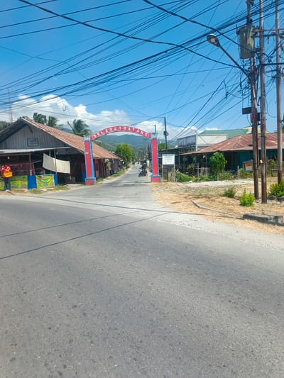 The image shows a street scene in Tenggela, Indonesia, on a clear day. An archway with the Indonesian greeting "SELAMAT DATANG" (Welcome) spans the road. To the left of the arch, a building with a wooden facade and a corrugated metal roof, labeled "PASAR SOURU", is visible. A person wearing an orange shirt and carrying a yellow basket stands in front of this building. To the right of the arch, several buildings with green and rusty metal roofs line the street. Overhead, a dense network of electrical wires crisscrosses the sky, supported by utility poles. A street light is visible on one of the poles. The road surface is asphalt, and the surrounding environment is dry with patches of grass and small bushes. In the background, a mountainous landscape can be seen under the blue sky. A motorcycle is passing through the archway.