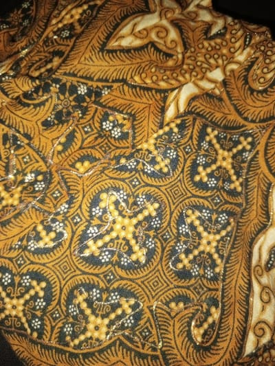 The image displays a detailed, close-up view of a textile fabric, located in Na Sang, Laos. The fabric is adorned with a complex, multi-colored pattern featuring dominant shades of dark gold/ochre, dark blue-black, and off-white/cream. The design incorporates intricate curvilinear motifs, including stylized foliage and scrolls, alongside repeating geometric and floral elements. Specific patterns observed include small white four-petal flowers with yellow centers, and stylized gold cross-like shapes, often enclosed within circular and square frames against the dark blue-black sections. The textile is embellished with irregular, shimmering lines of metallic thread, likely gold, which highlight the contours of various design elements, particularly the cross-like motifs. The fabric exhibits minor creases, suggesting it is draped or folded, with an indistinct dark background visible. No people, actions, or specific environmental features beyond the fabric itself are discernible.