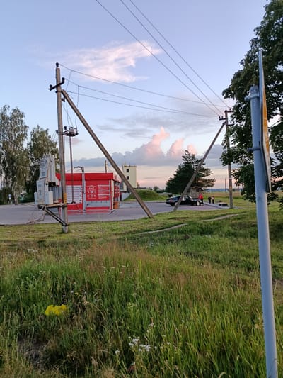 This is a daytime, medium-angle, full outdoor shot taken in the vicinity of Смілавіцкі сельскі Савет, Belarus. The scene depicts a rural roadside area with a car wash facility, a transformer, and overhead power lines. In the foreground, tall green grass and wildflowers dominate the landscape. A yellow plastic bag is visible in the grass on the left side of the frame.
In the middle ground, a red and gray self-service car wash facility with "24/7" signage is prominent. To the left of the car wash, a large electrical transformer is mounted on a concrete pole structure. Overhead power lines crisscross the sky, supported by two angled concrete poles, one of which appears damaged or unstable, leaning significantly. A black car is parked in the vicinity of the car wash, with its trunk open, and three individuals, two adults and one child, are near the car.
In the background, several trees, buildings, and more power poles are visible under a sky with scattered clouds, some tinged with pink, suggesting it may be late afternoon or early evening. A yellow and blue signpost is visible on the far right, partially obscuring the view of a large tree. The overall environment appears to be a semi-rural or suburban area with infrastructure for both services and utilities.