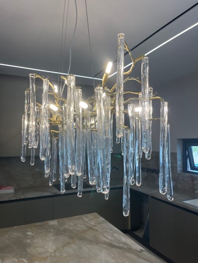 The image shows a modern chandelier with clear, icicle-like glass droplets hanging from a gold-colored, branch-like frame. The chandelier is illuminated by several small, cylindrical LED bulbs. It is suspended from a white ceiling with two linear LED light strips. Below the chandelier, a kitchen area is visible, featuring dark gray cabinetry, a marble countertop, and a backsplash with a patterned design. A window with a dark frame is partially visible on the right. The location is identified as Moni Community, Cyprus. There are no people or animals present in the image.