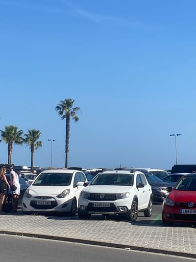 The image depicts a parking lot in Conil De La Frontera, Spain, under a clear blue sky. Several cars are parked, including a white Hyundai and a white Dacia Sandero, with a red Volkswagen Golf to the right. Palm trees are visible in the background, suggesting a coastal or warm climate. A few people are present near the cars, one of whom is standing next to a bicycle. The ground is paved with interlocking bricks.