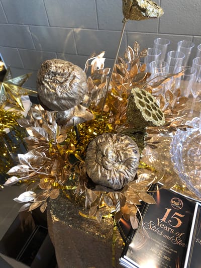 This is a close-up, high-angle shot of a decorative arrangement featuring gold-colored artificial leaves, lotus seed pods, and spherical ornaments that resemble dried gourds or ammonites. The decor is set against a backdrop that includes a tiled wall on the left and a stack of black programs with gold lettering on the right. The programs advertise "15 Years of Stars and Stories," an "Anniversary Gala Concert and Celebration" for "Star School," scheduled for "June 26, 2026." Scattered gold sequins and glitter add to the festive, celebratory theme. The overall impression is one of a special occasion, possibly a school anniversary event, with opulent decorations.
