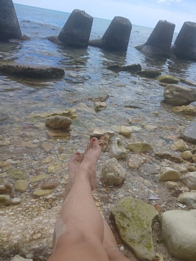The image shows a close-up of a person's legs and feet on a rocky beach with the sea in the background. The person's feet are bare, with red painted toenails, and are partially submerged in shallow, clear water. The beach is covered in small to medium-sized stones and pebbles, some of which are covered in algae. In the background, there are large, concrete breakwaters rising from the water, creating a barrier against the sea. The sea itself is calm with gentle waves. The sky is clear and blue, suggesting a sunny day. The scene is located on the coast near the village of Osenovo, Bulgaria.