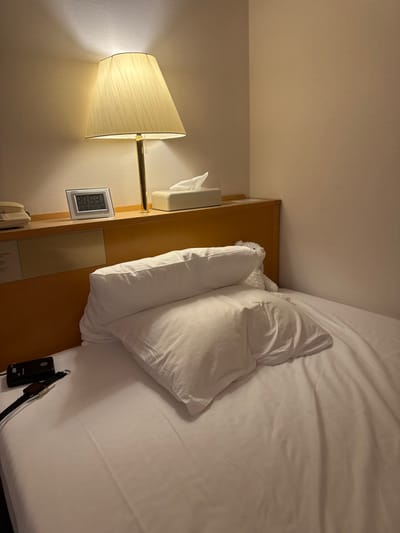 The image displays an unoccupied single bed within a room, characteristic of a hotel or similar accommodation, situated in Toyota, Japan. The bed is furnished with white sheets and two white pillows; one is propped upright against a light-colored wooden headboard, while the other rests horizontally in front of it.
On the wooden shelf above the headboard, from left to right, are: a beige landline telephone receiver, a silver-framed digital alarm clock displaying "20:53," "11/20 Fri," and "23.8°C," a brass-stemmed table lamp with an illuminated pleated off-white fabric shade, and a rectangular box of white tissues. On the left side of the bed, a black electronic device, possibly labeled "MB2," with a white cable attached, lies beside a dark-colored watch. The room features plain light-colored walls, with the corner of the room visible to the right. The primary light source is the illuminated bedside lamp. No individuals or specific actions are depicted.