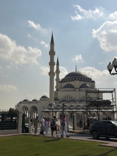 This image depicts the Sharjah Mosque in Sharjah, United Arab Emirates, under a partially cloudy, bright daytime sky. The large, cream-colored mosque features a prominent central dome, numerous smaller domes, and two tall minarets with dark grey conical tops.
In the foreground, a manicured green lawn is visible on the left. A paved pedestrian pathway or road extends across the mid-ground. Approximately eight to ten individuals are walking from left to right along this paved area. Their attire is mixed, with several men wearing traditional light-colored thobes and head coverings (ghutras or keffiyehs), while others are dressed in modern casual wear including t-shirts, shorts, and sneakers. One individual wears a backpack.
To the right of the walking figures, a dark-colored SUV is partially visible, positioned on the paved surface, facing left. An orange and white traffic cone stands nearby. Further right, a decorative street light with multiple lamps is visible. A metal scaffolding structure, partially obscured by dark coverings, stands in front of the mosque on the right side. A dark metal fence delineates the green lawn from the paved area on the left, behind which another dark vehicle is partially visible. Additional individuals are observed near the mosque's arched entrance structures, some appearing to be seated.