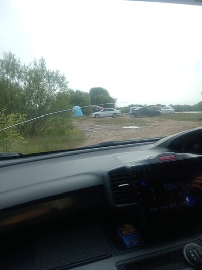 The image shows a view from inside a car parked on a dirt road. The sky is overcast. To the left, there are trees and grass. In the background, there are several parked cars, a blue tent, and other tents. The ground appears wet and muddy, with puddles. The car's dashboard is visible in the foreground, with a radio display showing the frequency "106.10 MHz". A pack of cigarettes is on the dashboard. The location is identified as Vasil'evka, Russia.