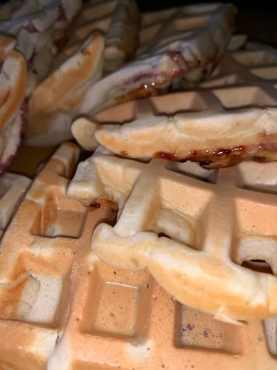 The image displays a close-up, overhead view of several golden-brown waffles, some of which are layered on top of each other. The waffles have the characteristic grid pattern with indentations. There are streaks of a dark red, viscous substance, likely jam or fruit preserve, visible in the crevices and on the surface of some of the waffles, particularly on the left side of the frame. The lighting is dim, creating a slightly shadowed effect and highlighting the texture of the waffles. The background is dark and out of focus. The image does not contain any people or visible text. The scene appears to be a close-up of a food item, likely prepared for consumption, in a domestic setting given the nature of the food.