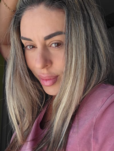 A close-up, slightly angled, medium-shot photograph shows a woman with long, highlighted blonde hair. She is looking towards the camera with a neutral expression. Her eyes are brown, and her eyebrows are dark and well-defined. Her lips are full and appear to be wearing a subtle pink lipstick. She is wearing a pink t-shirt. The background is dark and out of focus. There is no information in the image to suggest the location is the city of Carpi, Italy.
