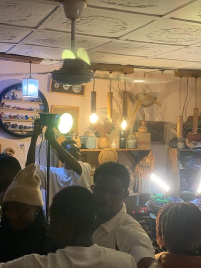 The image shows the interior of a shop in Ita Oniyan, Nigeria, located at the coordinates 7.282 latitude and 5.161 longitude. There are four people in the room, two of whom are standing in front of a shelving unit. One of the standing people is holding a large, white, circular light source, while the other stands with their back to the camera and appears to be setting up equipment. Another person, sitting with their back to the camera, is wearing a light tan hat. The fourth person is sitting in the foreground, facing the camera. All four people are Black and appear to be African. The room is lit by three overhead lights, one large, white, circular light source, and three small, white, rectangular lights. The room has white walls with a textured pattern. The ceiling has a fan. The shelving unit has a variety of objects on it, including a bottle, a glass jar, a vase, a small white ceramic container, and several other objects. The room has a wood floor.