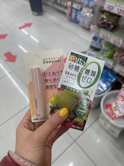 A person's right hand is holding two packages in a store. The hand has yellow nail polish on the index finger. The packages are both for food products. The package on the left is for a "deeper" brand lip balm. The package on the right is for "deep-ut" brand green tea cookies. The person is standing in a store, likely a convenience store, as the floor is tiled and there are shelves in the background. The store is in the Minato City area of Tokyo, Japan. The coordinates provided place the store in a commercial district. The shelves in the background are stocked with various goods, likely snacks and drinks. The lighting is fluorescent, and the overall environment is clean and well-lit.
