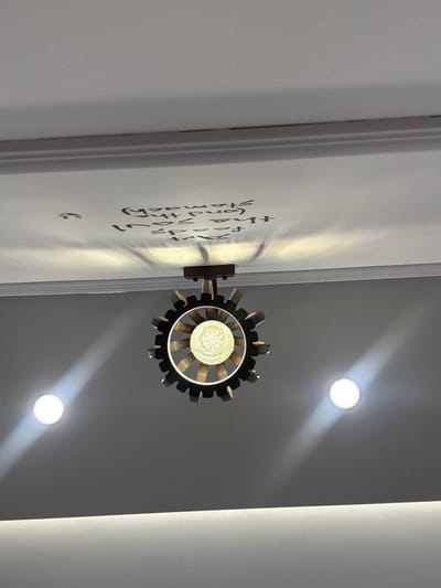 A low-angle shot shows a wall-mounted light fixture shaped like a gear. The fixture emits a warm glow. On the wall, near the fixture, there is some writing. Above the light fixture, there are two ceiling lights emitting bright light beams. The walls are primarily white and gray. The perspective is slightly off-center.