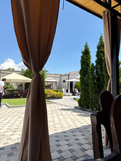 The image captures an outdoor leisure area in Jrvej, Armenia, viewed from under a brown canopy with curtains tied back on both sides. In the immediate foreground, a patterned paving stone path covers the ground, featuring interlocking light and dark gray rectangular blocks. On the left, a vibrant green artificial turf area leads to a beige, square-shaped gazebo with "Coca-Cola" branding on its canopy, under which wooden picnic tables and benches are arranged. Several individuals are present near the gazebo, including a man in a white t-shirt and dark pants walking left, partially obscured. In the right mid-ground, a line of tall, slender, dark green coniferous trees stands beside a light-blue pool or circular water feature, accompanied by white sun loungers and a white patio umbrella. A white building with a visible roof section is located behind this area. The background features a light-colored wall and another, smaller white gazebo structure. The sky is clear blue with scattered white cumulus clouds, indicating bright daylight and casting distinct shadows on the paving stones.
