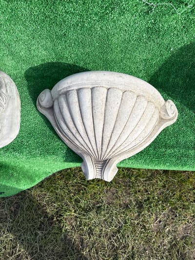 The image shows a grey, shell-shaped architectural ornament, possibly made of concrete or stone, resting on a square of artificial green turf. To the right of the turf, there is a patch of bare earth with sparse vegetation. The photograph was likely taken outdoors in daylight. The location in the city of Chirk, United Kingdom is not discernible from the image. There are no people visible in the image.