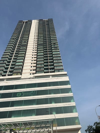 An eye-level, exterior, wide-angle shot captures the Kingsgate International School building in Subang Jaya City Council, Malaysia, under a clear blue sky. The building is a tall, modern high-rise with a distinct architectural style. The lower portion of the building features large, reflective green glass windows, suggesting a commercial or institutional use. A crest and the words "KINGSGATE INTERNATIONAL SCHOOL" are visible on these windows. Above this section, the building transitions to a residential-looking facade with numerous balconies and windows, arranged in a repeating pattern. The building's structure ascends uniformly, with a central lighter-colored section flanked by darker balcony areas. The shot is angled upwards, emphasizing the height of the building against the sky. A street lamp and some trees are visible on the right side of the frame, indicating the building's urban or suburban setting. The image is well-lit, with shadows indicating a bright, sunny day.