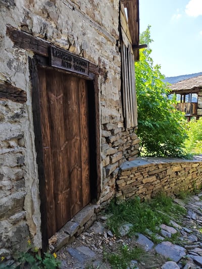 This image depicts the exterior of a historic building in the village of Kovachevitsa, Bulgaria. The central focus is a rustic, dark brown wooden door with vertical planks, featuring a simple metal latch on its right side. Above the door, a dark metal or wooden sign is affixed to a prominent wooden beam, bearing raised Cyrillic text: "КИЛИЙНО УЧИЛИЩЕ ЛЕТО 1820ТО." This translates to "CELL SCHOOL YEAR 1820," indicating the building's historical function.
The building's walls are constructed from irregular, light-colored stones, with some sections exhibiting a rough plaster finish and exposed dark wooden structural beams. To the immediate right of the door, the stone wall continues, and a narrow, vertically planked wooden structure is visible, likely part of a window or architectural extension.
In the foreground, a cobbled pathway of uneven stone slabs is partially covered with patches of green grass and a few small yellow wildflowers. A low, dry-stacked stone wall extends from the right side of the building, delineating the pathway. Beyond this wall, vibrant green foliage from a tree or large bush is prominent, partially obscuring the view. In the background, beneath a bright blue sky with scattered white clouds, other traditional village houses with distinctive brown-tiled roofs are visible, set against a backdrop of distant green mountains. There are no people present in the scene.