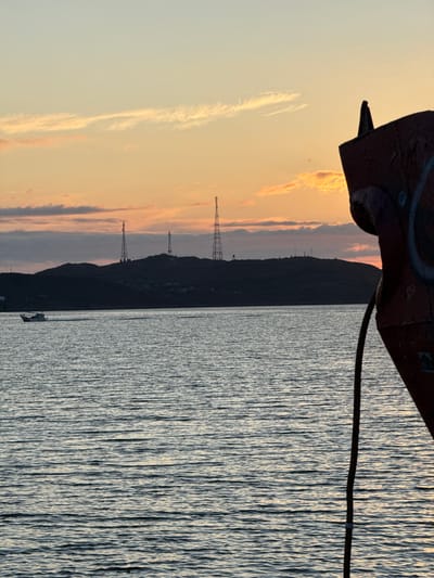 This image depicts a view of the Teriberka Bay in Teriberka, Russia, during sunset. The sky is a gradient of pale blue and orange, with wispy clouds. In the distance, a silhouetted hill is visible, topped with communication towers and some buildings. A small white boat is moving across the water. In the foreground, the rusted bow of a ship juts into the frame from the right, with a thick rope hanging down. The water is calm with ripples.