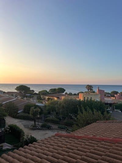 This is a horizontal, eye-level, medium shot of a coastal town at sunset. The sky transitions from a pale yellow-orange at the horizon to a clear, light blue overhead. The sea is visible in the background, calm and reflecting the sky's colors. In the middle ground, a cluster of buildings with terracotta tile roofs and pastel-colored walls are nestled among lush green trees. The architecture appears Mediterranean, with some buildings featuring arches and balconies. A narrow road winds through the settlement, and two bicycles are parked near a lamppost on a gravel path. The foreground is dominated by a close-up view of a tiled roof, its weathered terracotta tiles showing the texture and color variations of age. The scene conveys a peaceful, serene atmosphere, characteristic of a seaside village in the late afternoon or early evening. The location is identified as Biddeputzi/Villaputzu, Italy.
