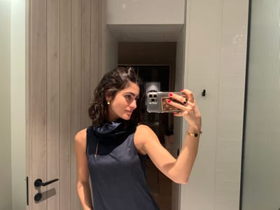 A woman with curly dark hair wearing a sleeveless dark blue dress with a high, draped neckline takes a mirror selfie. She holds her phone up with her right hand, showing a Louis Vuitton patterned phone case and red nail polish. She has a gold bracelet on her right wrist and gold stud earrings. The background shows a bathroom with a wooden door on the left and a tiled wall and shower door on the right.