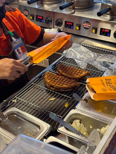 The scene depicts an indoor food preparation area at a Famous Belgian Waffles outlet in Panglao, Philippines. In the foreground, an individual, identified as an employee by an ID badge bearing the name "ABBIGAIL C. MACUJA" and wearing an orange and brown uniform with a black face mask, is actively using metal tongs. They are placing a prepared, semi-circular waffle into an orange paper sleeve, which is part of the brand's packaging.

On a metal cooling rack in the center, two additional semi-circular waffles, filled with a red jam or fruit spread and a white cream-like filling, are visible. Another partially packaged waffle rests within an orange cardboard box labeled "Famous BELGIAN WAFFLE" on the right side of the rack. Scattered crumbs and small food debris are present on the rack below the waffles.

Adjacent to the rack, several stainless steel condiment containers are arranged. One holds a quantity of white, granular substance (possibly grated cheese or a sugar topping) with a metal spatula, while another contains a dark, viscous liquid.

In the background, three stainless steel commercial waffle machines are visible. Each machine features digital displays showing numerical values (0303, 0339, 0329) and control knobs. One machine is marked with a "Famous BELGIAN Waffles" sticker. The overall environment suggests a bustling, functional commercial kitchen or food service counter.