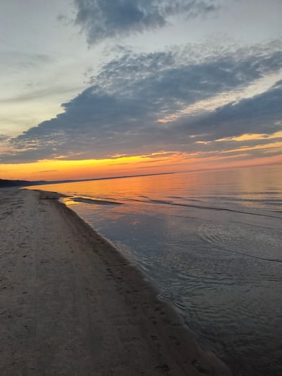 A wide-angle, vertical shot captures a beach and sea at sunset in Apšuciems, Latvia. The foreground is dominated by a sandy beach with visible footprints, curving to the left and disappearing into the distance. The sea, to the right, is calm with gentle ripples reflecting the warm hues of the sunset. The horizon is marked by a distant, dark landmass. Above, the sky transitions from a bright orange and yellow band at the horizon, indicating the setting sun, to soft pinks and grays, with dark, textured clouds scattered across the upper portion of the frame. The overall impression is one of serene natural beauty.