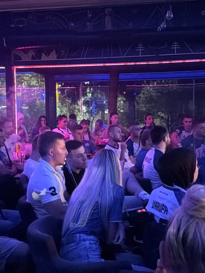 The image depicts a group of people gathered in what appears to be an indoor smoking lounge or cafe, possibly in Travnik, Bosnia and Herzegovina, given the context. The atmosphere is dimly lit with vibrant neon and LED lighting, primarily in shades of pink and blue, casting a colorful glow on the patrons and the surroundings. Many individuals are seated, some appearing to be smoking shisha pipes, indicated by the presence of hoses and smoke. The people are casually dressed, and some are wearing sports-like jerseys, one of which has the name "JARUPA" visible on the back. The environment includes seating arrangements, tables with items on them, and decorative elements on the walls and ceiling. The overall scene suggests a social gathering in a relaxed, potentially entertainment-oriented venue.