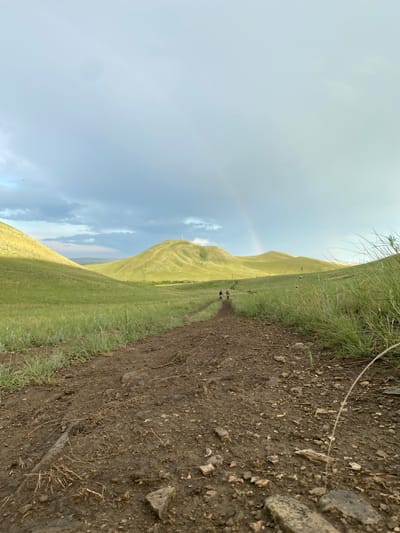 In the city of Verkhneozernoye, Russia, a dirt path winds through rolling green hills under a cloudy sky. A faint rainbow arcs across the horizon, suggesting recent rainfall. Several figures are visible walking along the path in the distance, heading towards a prominent, rounded hill topped with a structure. The foreground of the path is muddy and strewn with small rocks and dry vegetation. The grassy slopes on either side of the path are tall and appear somewhat unkempt.