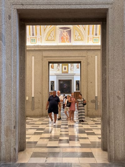 In San Lorenzo De El Escorial, Spain, within a grand hallway of what appears to be a historical building or museum, the image captures a view through a large stone archway. The hallway features a black and white checkered marble floor that creates a visually striking pattern. Several individuals are walking through the hallway, some towards the viewer and others away.

In the foreground, to the right and left, are large, textured stone columns framing the entrance. Beyond the archway, the hallway opens up to reveal more of the building's interior. Prominently displayed on the walls are various framed artworks, including paintings and decorative murals, suggesting an art gallery or a historically significant location. The ceiling area above the hallway is adorned with intricate murals featuring figures and architectural elements, illuminated by soft lighting.

The people visible in the scene are casually dressed, indicating they are likely visitors. One man in the foreground is wearing a dark blue polo shirt and dark shorts, walking towards the camera. To his right, a man in a white shirt and beige pants is also walking in the same direction. Further into the hallway, a group of three women is walking away from the camera. One woman in the center wears a pink dress, and another to her right is in a black and white striped dress. The overall atmosphere suggests a leisurely visit to a place of cultural and artistic significance.
