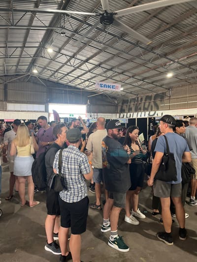 The image shows a large gathering of people in a large, open, industrial space.  The space appears to be a warehouse or large shed. There are large, metal support beams overhead and exposed metal sheeting walls, along with a large, rotating fan.  The space has a corrugated metal roof. The people appear to be socializing and drinking.  There are many tables and chairs arranged throughout the space. Several people appear to be in conversation while others are holding alcoholic beverages.  The people are dressed in casual clothing, many wearing t-shirts and shorts.  The space is located at the coordinates: -27.439, 153.075, which corresponds to a location within the city of Hamilton, Australia.  The address is in the industrial area of Hamilton North.  The image is likely taken at an event like a market or festival, and some people are wearing badges and lanyards.