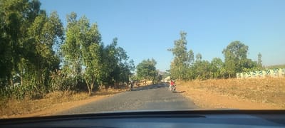 The image captures a straight, worn asphalt road under a clear blue sky, situated in a semi-rural area of Hoss, Nigeria. Two motorcycles are observed traveling away from the viewer on the road. One, a darker motorcycle, occupies the left side of the lane, while a lighter-colored motorcycle is positioned closer to the right shoulder. Each motorcycle appears to be carrying at least one occupant. The road is flanked by reddish-brown, dry earth. On the left, dense green foliage, including tall trees, lines the roadside. On the right, similar vegetation is present, along with a distant, partially visible structure featuring green and white patterns. The perspective suggests the photograph was taken from within a vehicle, with a dark dashboard element visible at the bottom of the frame.